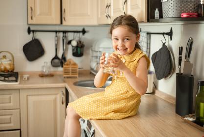 Petite fille souriante buvant dans un verre d’eau sur fond de cuisine.